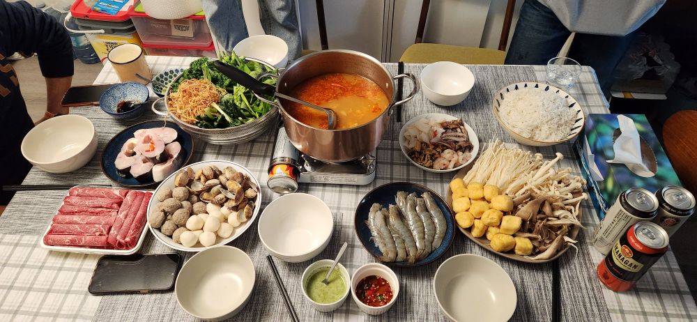 A picture of a table with a pot of tom yum soup base in the middle, surrounded by bowls of mushrooms, tofu, shrimp, clams, mussels, fish, sliced beef, vegetables, and rice noodles.