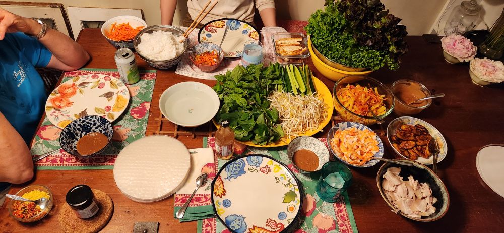 Numerous plates and bowls full of pork belly, shrimp, fried tofu, peanut sauce, rice paper, lettuce, cucumber, bean sprouts, pickled carrots, fried jicama, fried plantain, mint, fish mint, and Thai basil on a large wooden table.