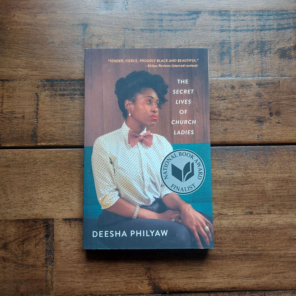 The book The Secret Lives of Church Ladies by Deesha Philyaw sits on a wooden surface. Cover is a photograph of a black woman with short hair wearing a collared shirt and bow time sitting on a teal bench.