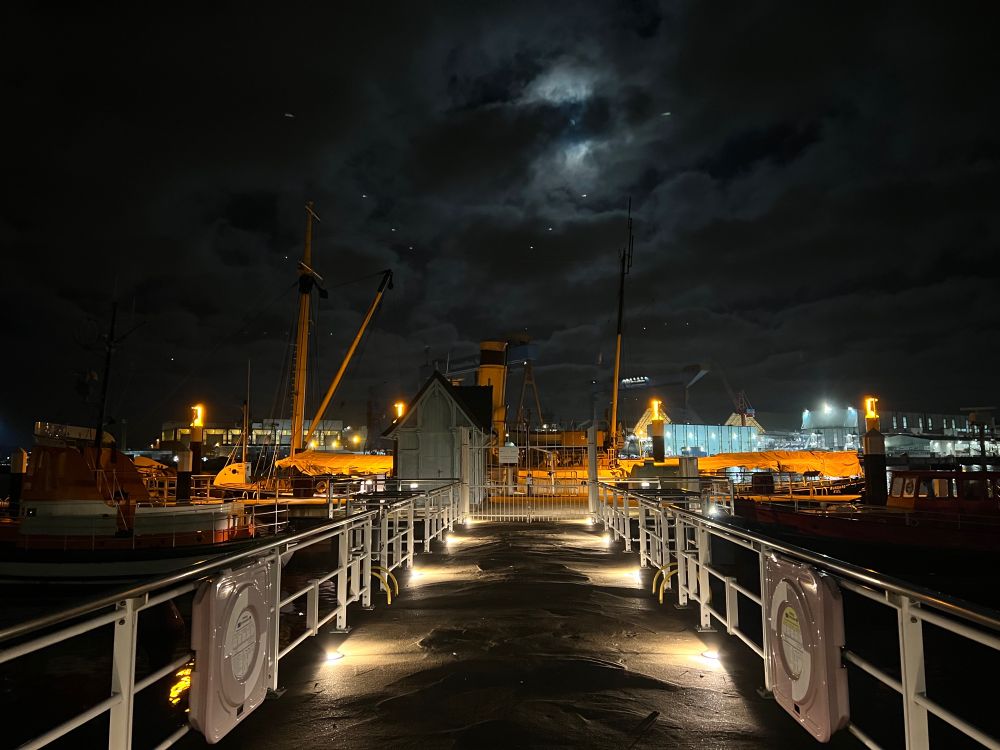 Ein Steg führt zu einem Dampfschiff. Es ist Nacht und der Vollmond ist schemenhaft hinter einer dünnen Wolkendecke zu erkennen.
