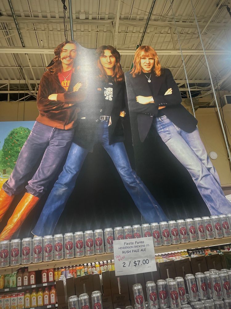 Cardboard cutout of the band Rush atop a shelf of Rush Beer