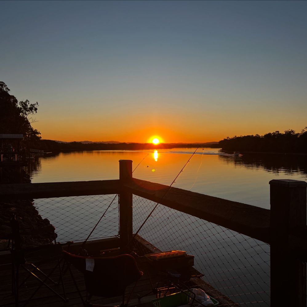 The photo is a shot of a corner of a wooden pier with a couple of fishing rods, camp chairs and other fishing gear overlooking wetlands, the sun is giving off an orange glow as it sets in the distance behind mangroves 