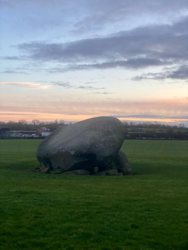 A dolmen which is a big rock stacked on smaller rocks surrounded by grass it’s sunrise and the sky is pastel blue and pink 