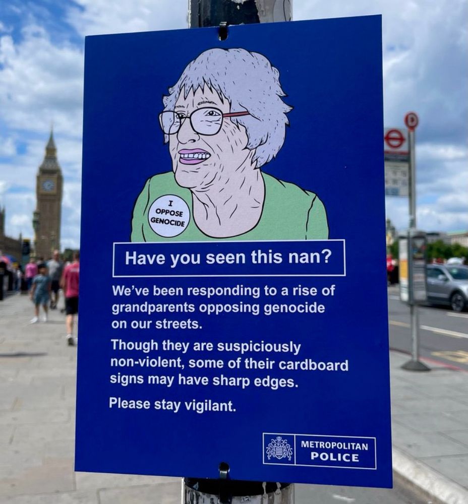 A parody sign posted on a London street near Big Ben shows an illustrated elderly woman wearing glasses and a badge that says “I OPPOSE GENOCIDE.” The text reads: “Have you seen this nan? We’ve been responding to a rise of grandparents opposing genocide on our streets. Though they are suspiciously non-violent, some of their cardboard signs may have sharp edges. Please stay vigilant.” The sign mimics official police warnings and is labeled with the “Metropolitan Police” logo