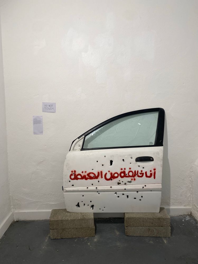A white car door, riddled with bullet holes, is displayed upright on two concrete blocks against a white wall. Red Arabic graffiti on the door reads “أنا خائفة من الحتمية” (translated: “I am afraid of the dark”). 