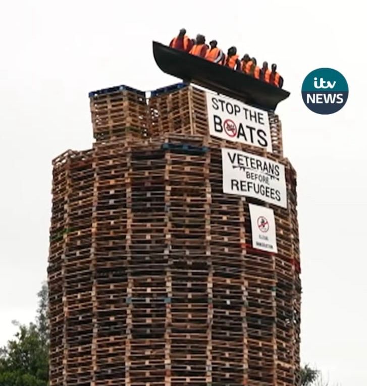 A large bonfire structure made from stacked wooden pallets stands in an open area, surrounded by fencing. On top of the pallets is a black model boat containing several human-shaped effigies wearing orange life jackets, resembling migrants. Attached to the pallets are banners reading “STOP THE BOATS” and “VETERANS BEFORE REFUGEES.”