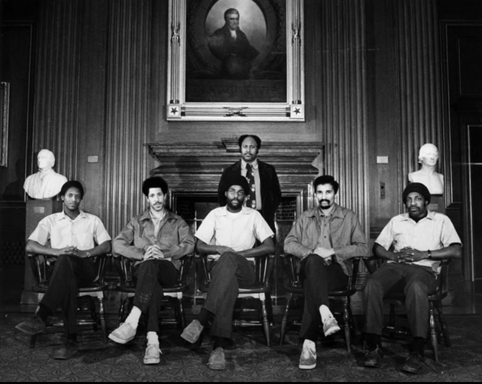 Black and white photo of six Black men sitting in the chairs of where the supreme courts official portraits are taken. They all look Regal 