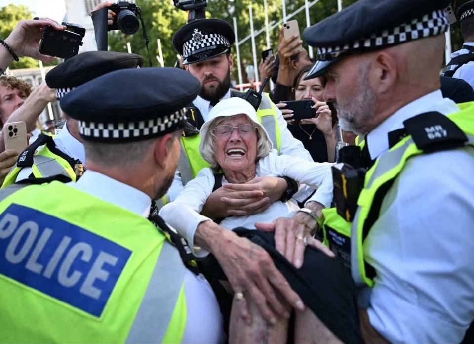 Policemen arresting an old woman. She’s had a grimace on her face and looks in pain. They are lifting her up 5 police officers 