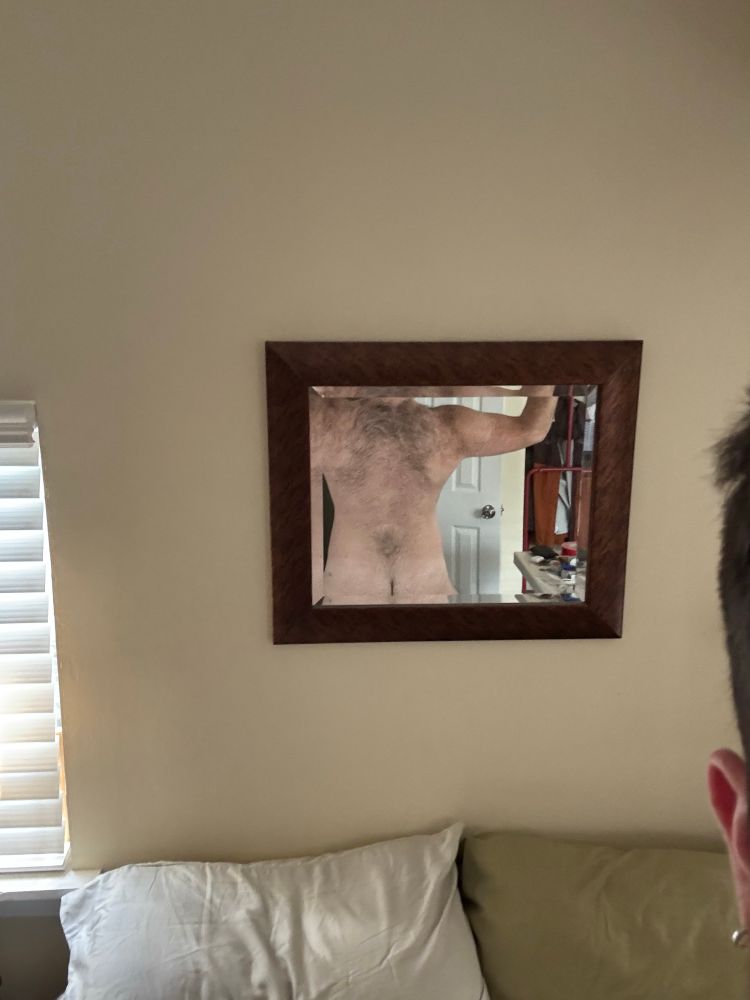 A somewhat artsy photo of a wall over a bed with a square framed mirror in the center, in the mirror is the back of a hairy white man showcasing the dark fur across his shoulders and upper back, the triangle of hair in the center of his lower back. He is flexing one arm and at the bottom of the picture is a hint of his pale ass. At the edge of the photo is an ear and a single silver hoop earring 