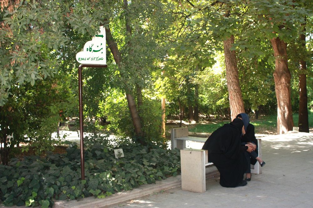 Photo of 2 women sitting on a bench surrounded by trees and plant beds. A sign reads "place of sitting." Bagh-e-Eram, Shiraz. Photo by me