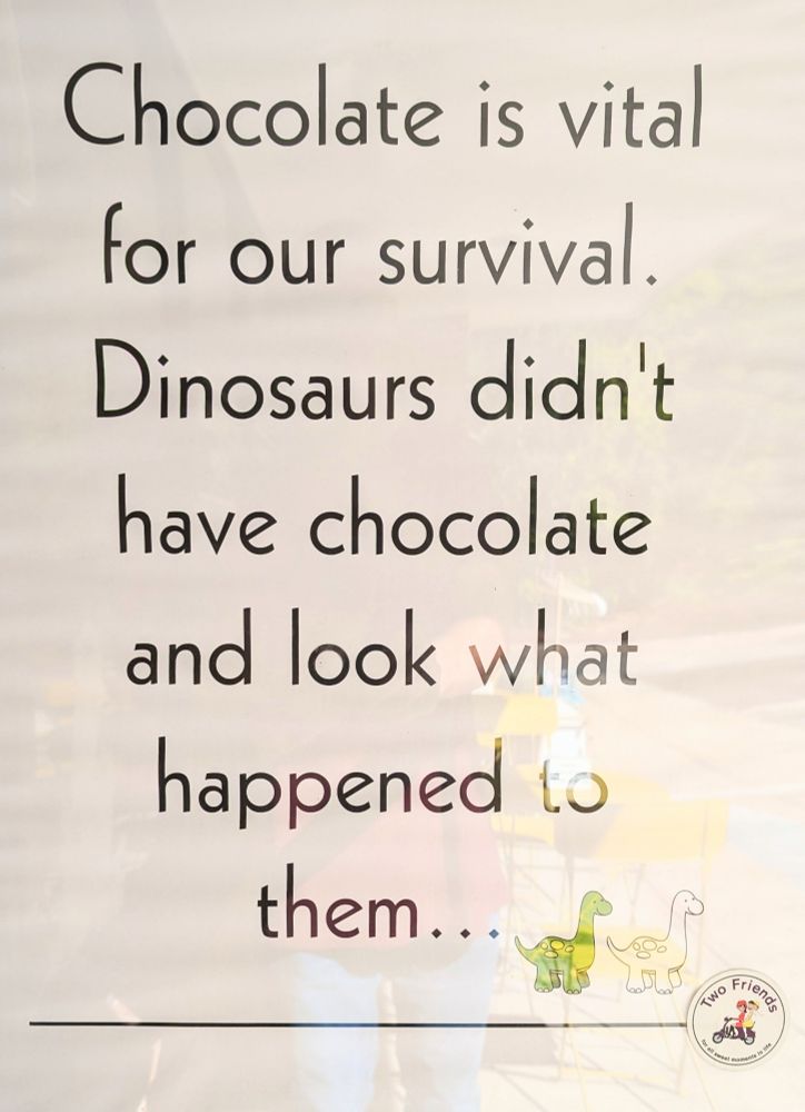 Photo of a sign outside Two Friends Chocolates, Boxborough Massachusetts. Sign reads "Chocolate is vital for our survival. Dinosaurs didn't have chocolate and look what happened to them...