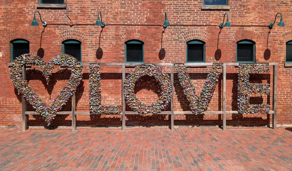 Photo of an art installation with the ❤️ symbol followed by "LOVE." The heart & each letter is made up of hundreds of multicolored padlocks. The installation is mounted on a wood frame in front of a brick wall which has 6 small arches windows in a row.