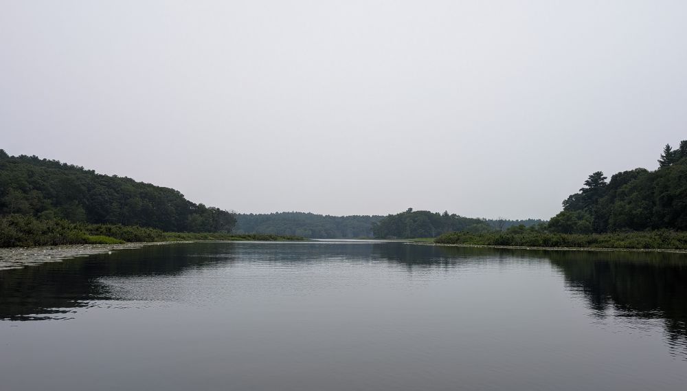 A wide, calm river stretches from foreground to the distance. Riverbanks are densely wooded as is the distant shore. The sky is a light gray haze from Canadian wildfires.