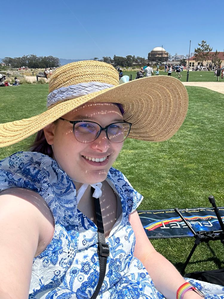 Selfie of me at the big lawn area of Presidio tunnel tops park. I’m wearing a blue and white sundress and a floppy straw hat with a blue band and bow. In the distant background is the palace of fine art building.