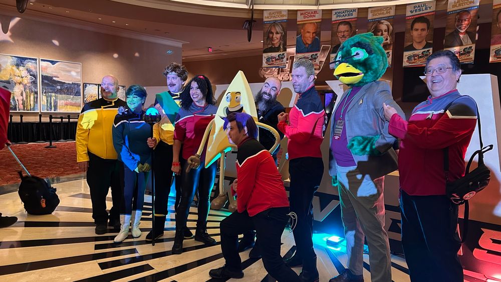Photo of named characters from Star Trek Lower Decks in front of the big STLV sign during the Lower Decks Cosplay Photoshoot. Pictured here from left to right cosplayers as: Shax, Tendi, King Billups, Badgey, Boimler, Ransom, Dr Migleemo (me!), and another Boimler (or Ransom not sure)