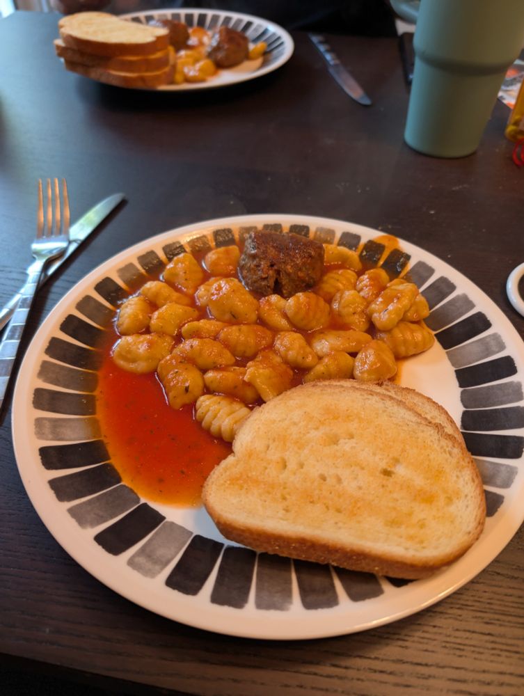 Plate with a large serving of gnocchi, smothered in homemade red sauce, with a meatball on top, and a side of garlic bread. 