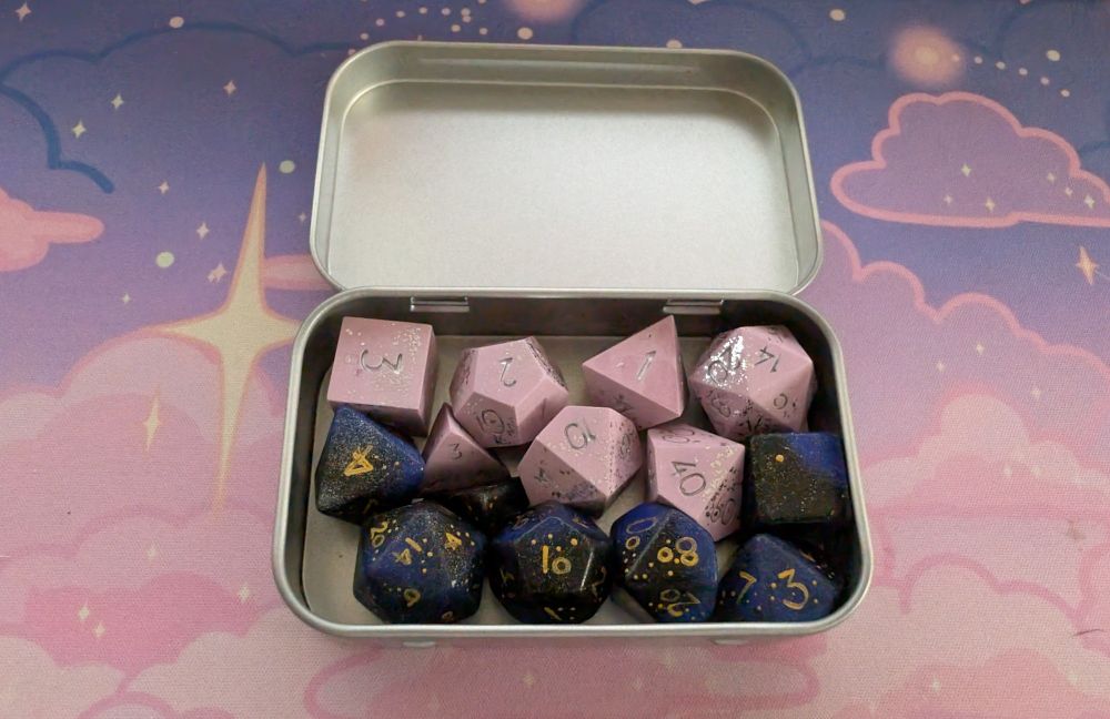Small silver rectangular tin holding two 7-piece dice sets. One is light pink stone with silver sparkles and silver numbers. The other is made of resin. The resin set is primarily dark purple, almost black, in color, with subtle medium purple swirled in, and gold painted numbers. 