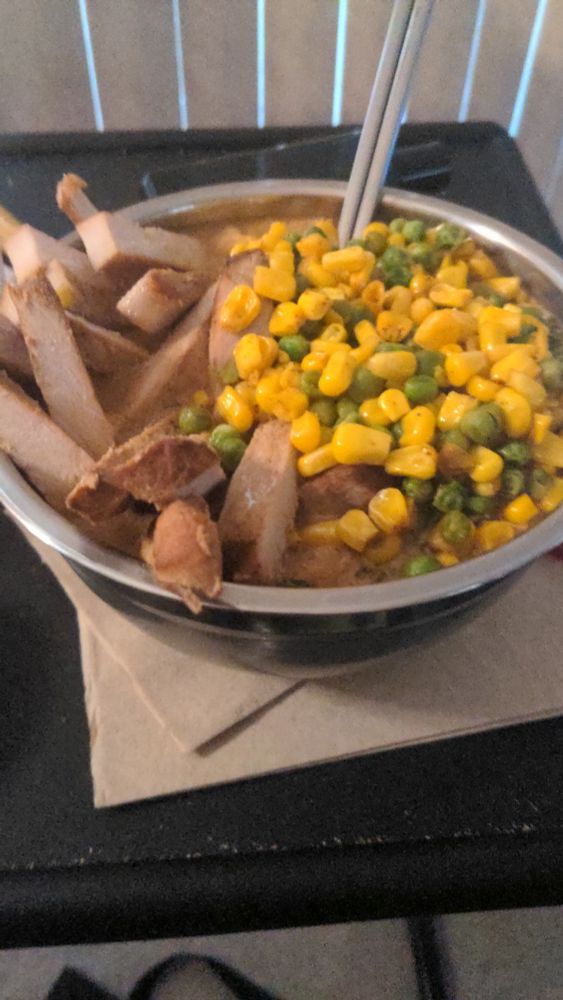 A metal bowl with Pork, Corn, Peas and broth covering a bed of noodles 
