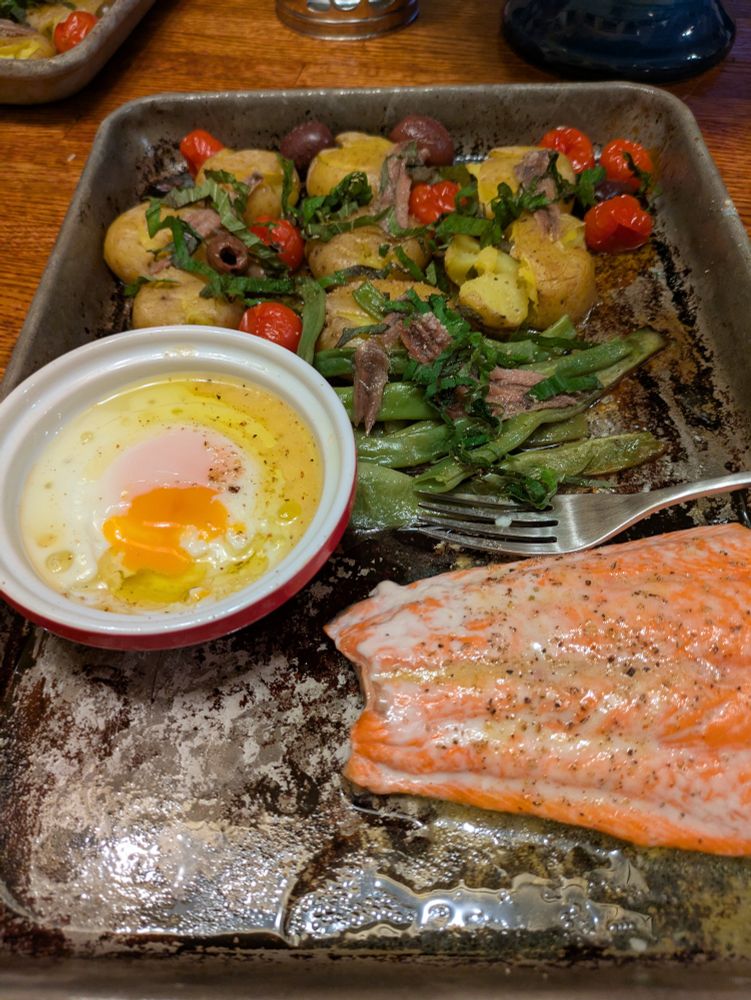 A sheet tray with an egg where the yolk is perfect, salmon, green beans, cherry tomatoes, olives, potatoes and anchovies. 