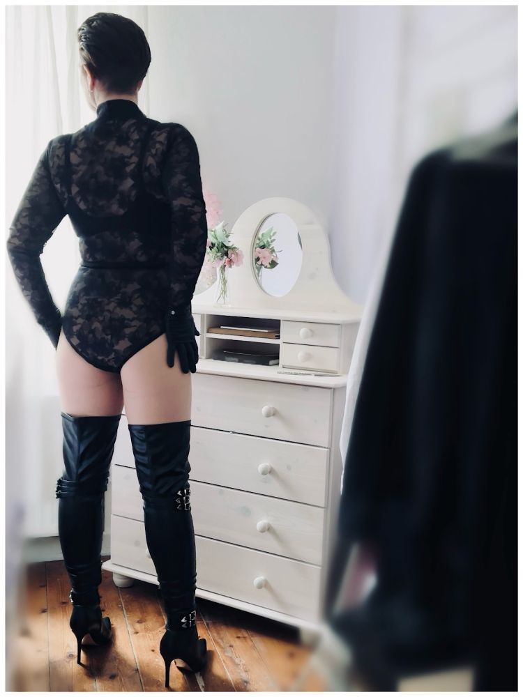 Skylla schaut aus dem Fenster und steht mit dem Rücken zur Kamera. Das Ambiente ist sehr hell, während Skylla in schwarz gekleidet ist. Sie trägt Overknee-Stiefel, Lederhandschuhe und einen Spitzenbody.