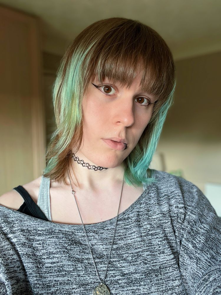 selfie of a white tgirl with blue and brown bangs and spiderbite piercings in a grey sweater