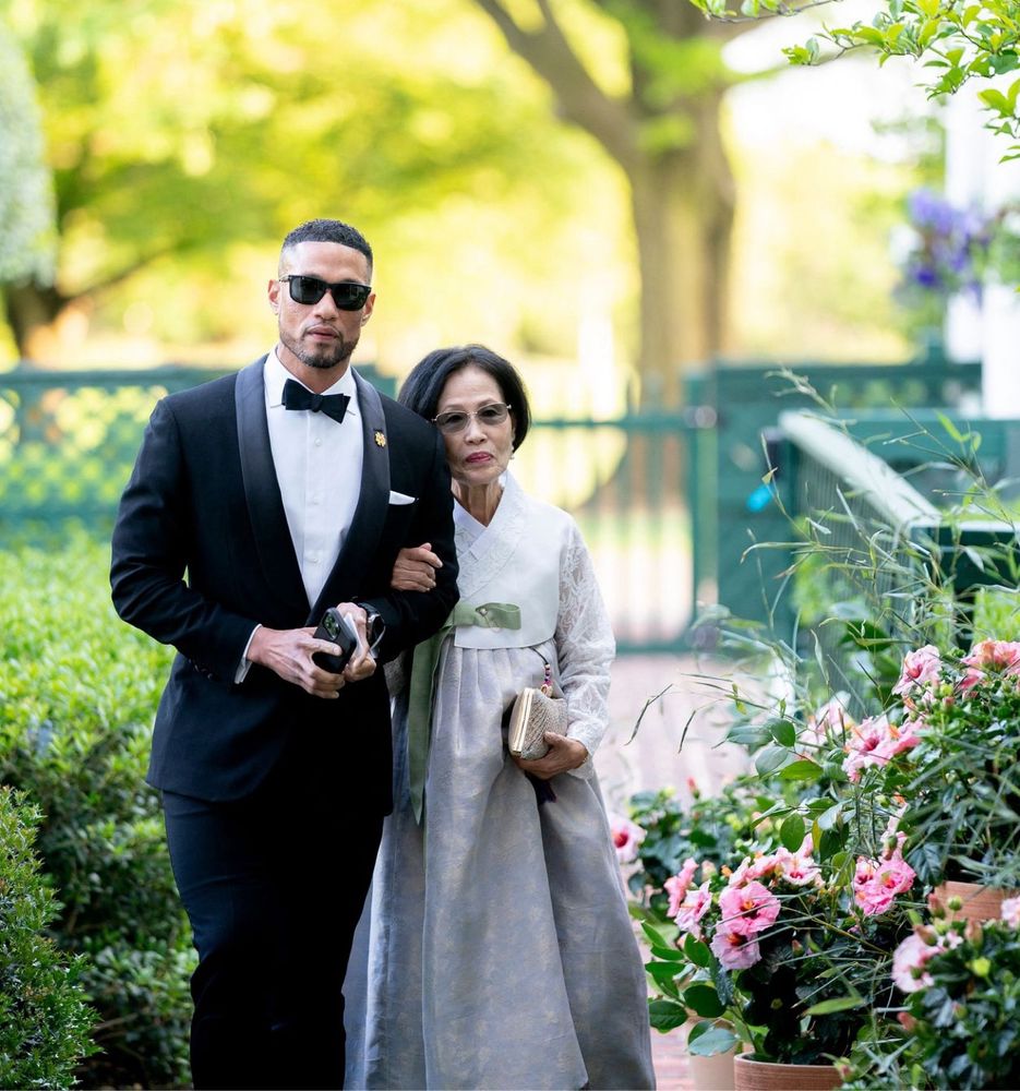 Image of the Norte Dame head football coach wearing a tux and his mother in traditional Korean formal dress in an outdoor garden setting