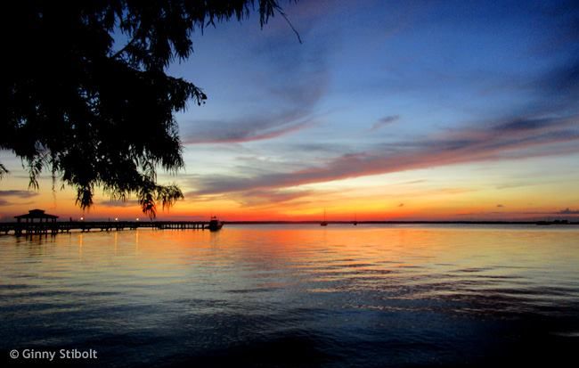 Photo of a pre-sunrise sky over a body of water. The sky is dark orange at the horizon, merging to a pale yellow and then from light blue merging to a dark blue. some wispy maroon-colored clouds are spread across the sky. The colors in the sky are reflected in the water. On the upper left side branches of a large tree are silhouetted in front of the sky. Also are the horizen on the left is a long deck exteending into the water.