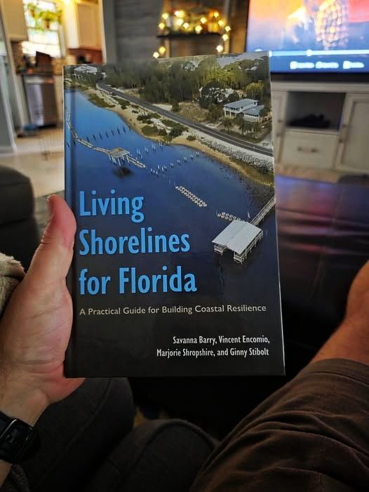 Photo of someone holding a book with a living room TV and kitchen out of focus in the background. The cover image is of a shoreline with vegetation and oyster reefs. Text says, "Living Shorelines for Florida A Practical Guide for Building Coastal Resilience Savanna Barry, Vincent Encomio, Marjorie Shropshire, and Ginny Stibolt"