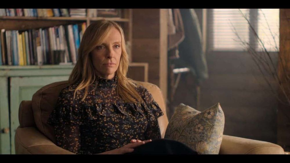 Joy Richards (Toni Collette) sitting in an light brown leather armchair in front of a bookcase. There is a throw pillow beside her on the chair covered in Morris & Co. “Strawberry Thief” in a light blue colorway. The birds and strawberries of the pattern aren’t visible, but green foliage and small cream and pink flowers can be seen.