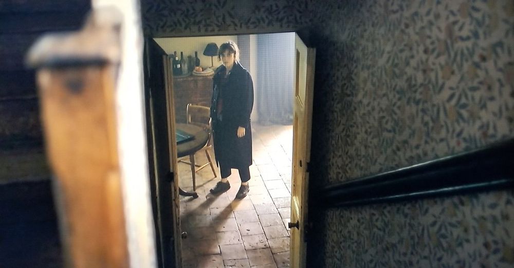 A view down a staircase and through a doorway into a room, where Marcella Backland (Anna Friel) stands looking up the stairs at the camera. The staircase is simple and enclosed by walls, which are papered in Morris & Co. "Fruit", a pattern of leaves, citrus, and pomegranates on a cream background. The room at bottom of the stairs has terra cotta tiles and simple wood furniture.