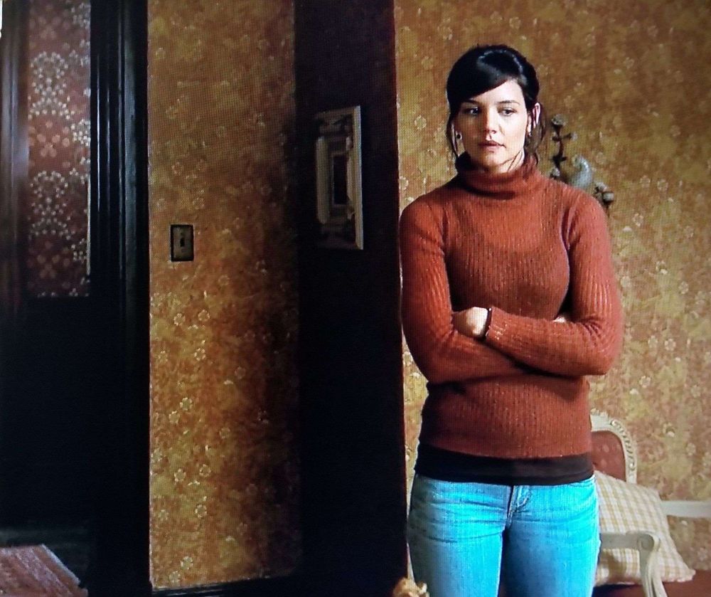 Kim Raphael (Katie Holmes) standing in a room papered with Walter Crane's "The Sleeping Beauty", a dense pattern of rose blooms and foliage with small figures intertwined, in shades of ochre gold. A dark paneled section of wall with Morris & Co. "Blackthorn" is visible at far left.
