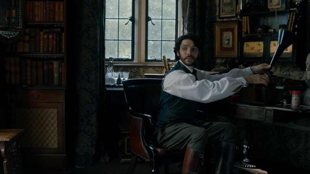 Nathan Appleby (Colin Morgan), a man with dark hair and beard in 1890s waistcoat, shirtsleeves, trousers, and high boots, sits at a desk adjusting a phonograph horn. Small amounts of “Indian” wallpaper are visible under the window and behind the various pieces of decoration above his desk.