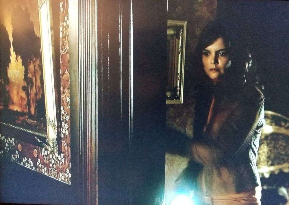 Kim Raphael (Katie Holmes) looking through a doorway trimmed with very dark wood. The wall on the side of the doorway has dark wood wainscoting topped by Morris & Co. “Blackthorn”, a symmetrical pattern of branches and small white flowers on a dark red background.