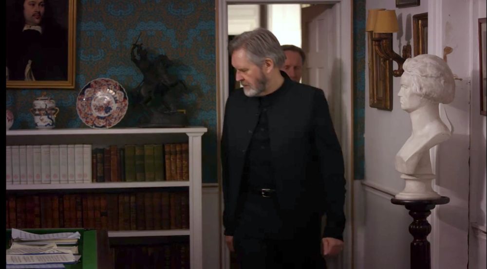 Setting: A room with low white wainscoting. To the right, the wall above the wainscoting is also white. To the left, taking up most of the frame, is a wall where the wainscoting is topped with Morris & Co. “Michaelmas Daisy”, a pattern of repeating symmetrical bunches of daisies with dense foliage. The colorway is dark bronze leaves and bright blue flowers on a dark blue background. There is a half-height bookshelf in front of the wall, full of antique books and toppes with a bronze of a horse and rider and some Chinese porcelain. There is a portrait of a man on the wall, maybe 17th-century. To the right, the white wall has other framed pictures on it and a marble bust. There is an open door in the corner where the two men meet, white with white trim.
Michael Falconer (James Fleet), a man dressed in black with grey hair and beard, is coming through the doorway into the room, followed by John Barnaby (Neil Dudgeon).