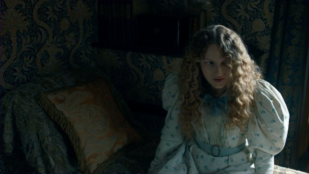 Harriet Denning (Tallulah Haddon), a young woman in a white and blue 1890s day dress with loose wavy blond hair, sits on a chaise lounge draped in fabric in front of a wall papered in Morris & Co. “Indian” wallpaper, a historic revival unrealistic pattern of very thick plant stems and large stylized flowers, in gold tones on a dark blue background.