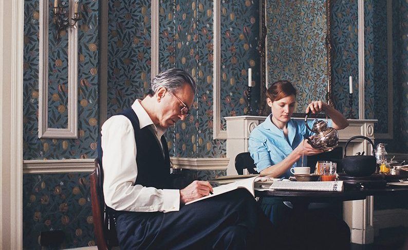 Setting: A room papered floor to ceiling with Morris & Co. "Fruit", a pattern of leaves, citrus, and pomegranates on a silvery blue metallic background. The room has white trim including a doorway, a chair rail, and inset panels above the chair rail; every divided section is papered with the same pattern. A large gilt-framed mirror sits on the mantle of a white fireplace, reflecting yet more wallpaper.
Reynolds Woodcock (Daniel Day-Lewis) and Alma Elson (Vicky Krieps) sit at a breakfast table in the middle of the room. Reynolds is writing or sketching in a notebook on his lap. Alma is pouring tea from a silver pot.