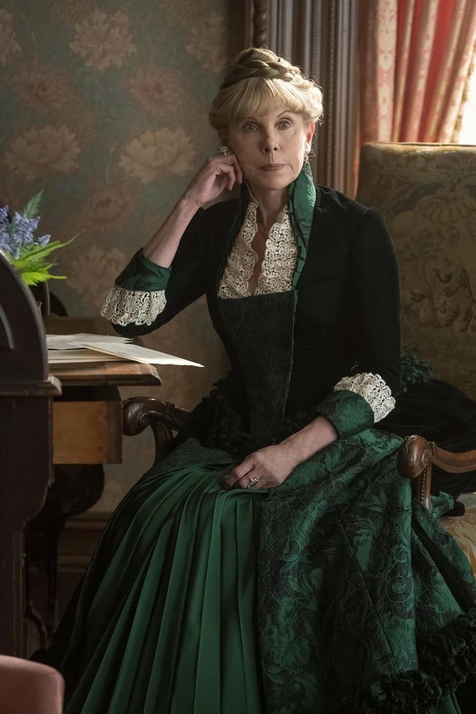 Agnes van Rhijn (Christine Baranski) sitting at a desk in the same room as the first image, with the wallpaper discussed behind her.