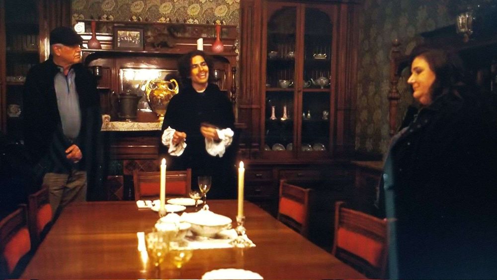 fills the middle of the room into the foreground, surrounded by low Aesthetic-style wooden chairs with small red back cushions. There is a limited setting on the bare wood table: two place settings at the far ends of the table, a small ceramic tureen, and two lit candles in low crystal candle sticks. against the walls are a mid-height buffet and two tall glass-fronted china cabinets. The buffet displays some pieces of Aesthetic pottery. The walls are papered in a green-on-green colorway of Morris & Co. “Chrysanthemum” (see second image for description). 

Three people stand around the table: an older man in casual modern clothing, a younger man with long dark hair and a dark jacket over a white shirt with big flowing white cuffs, and a woman in a dark coat or dress.