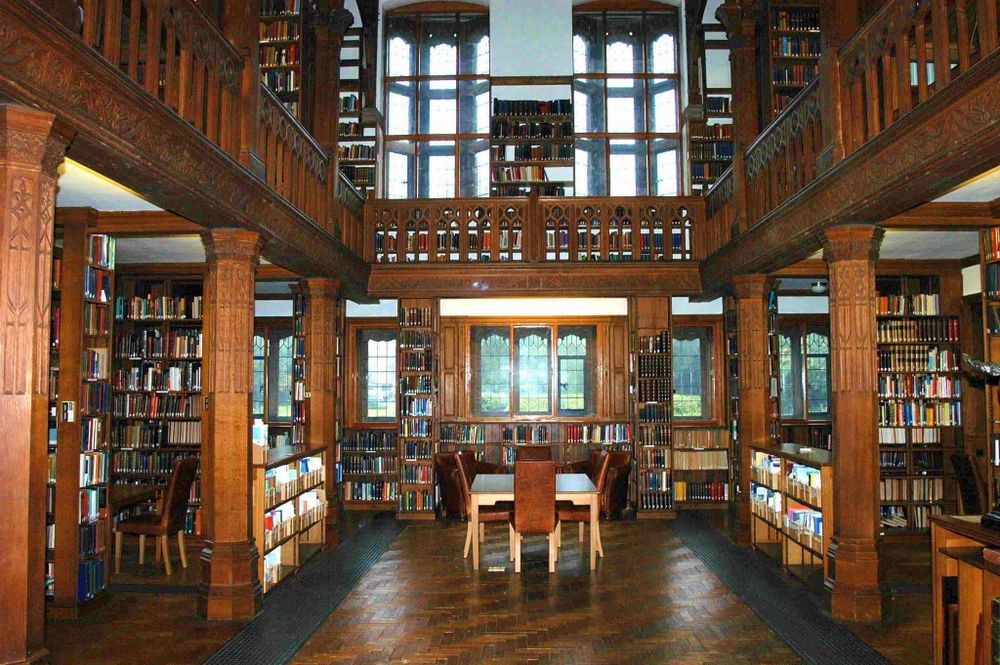 A photo of the Reading Rooms at Gladstone's Library, taken 16 years ago. The pillars, floors, walls and gallery are dark wood, with bookcases filling the space. A table with four chairs around it sits in the middle.