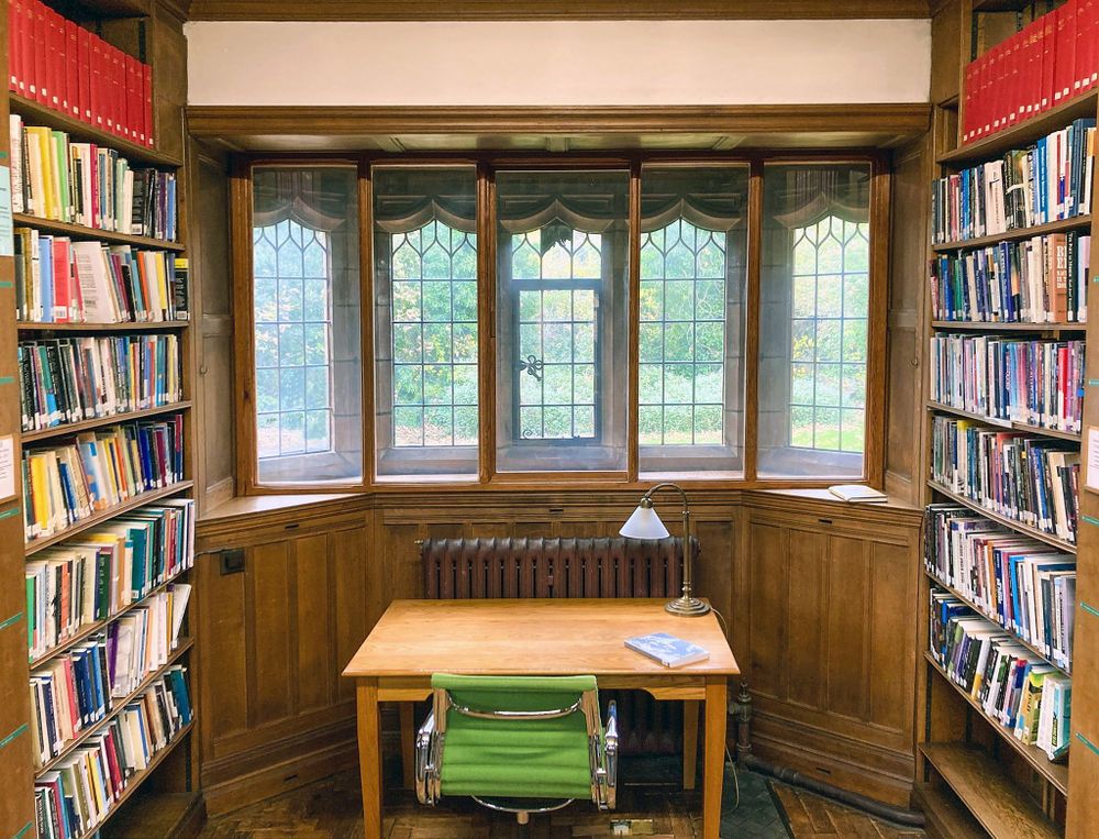 A desk in a space between bookshelves. The desk is set in front of a bank of leaded windows. There is a green chair in front of it and a lamp on the corner of the desk.