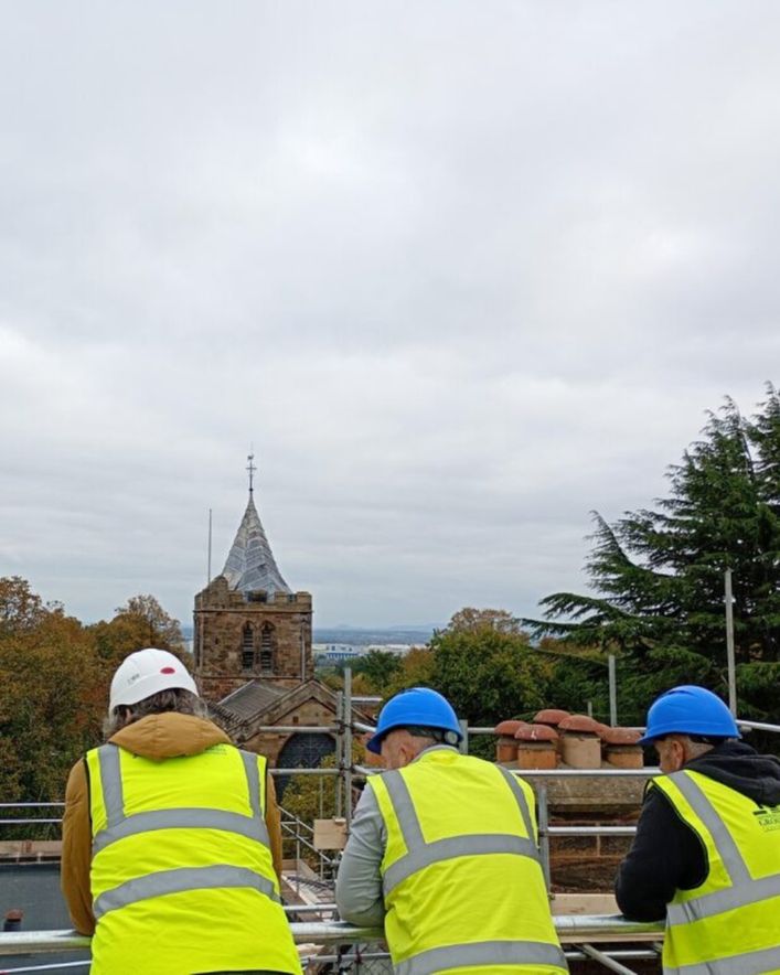 Three construction workers wearing high-visibility vests and hard hats stand on scaffolding with their backs to the camera, looking out toward an old stone church with a tall spire. Trees with autumn foliage surround the church, and the sky is overcast.
