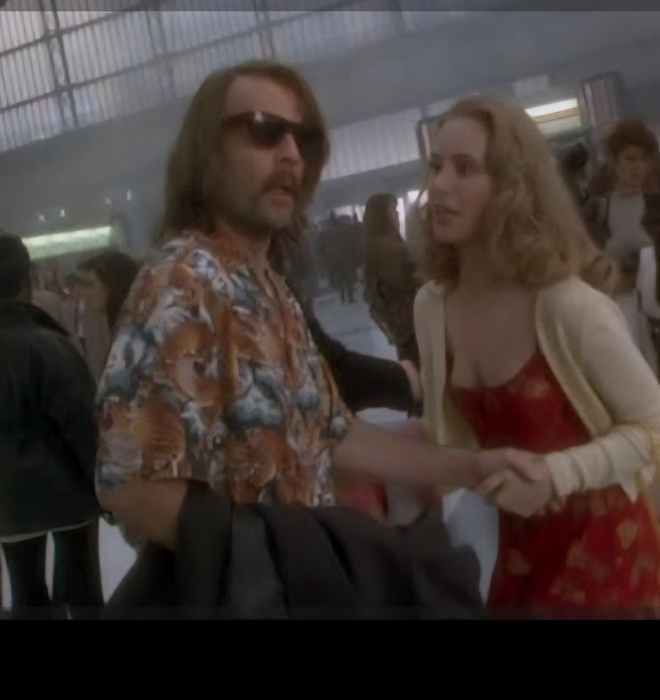 Cena do filme Os 12 Macacos Homem branco de óculos e bigode falso, mulher branca de peruca loira.