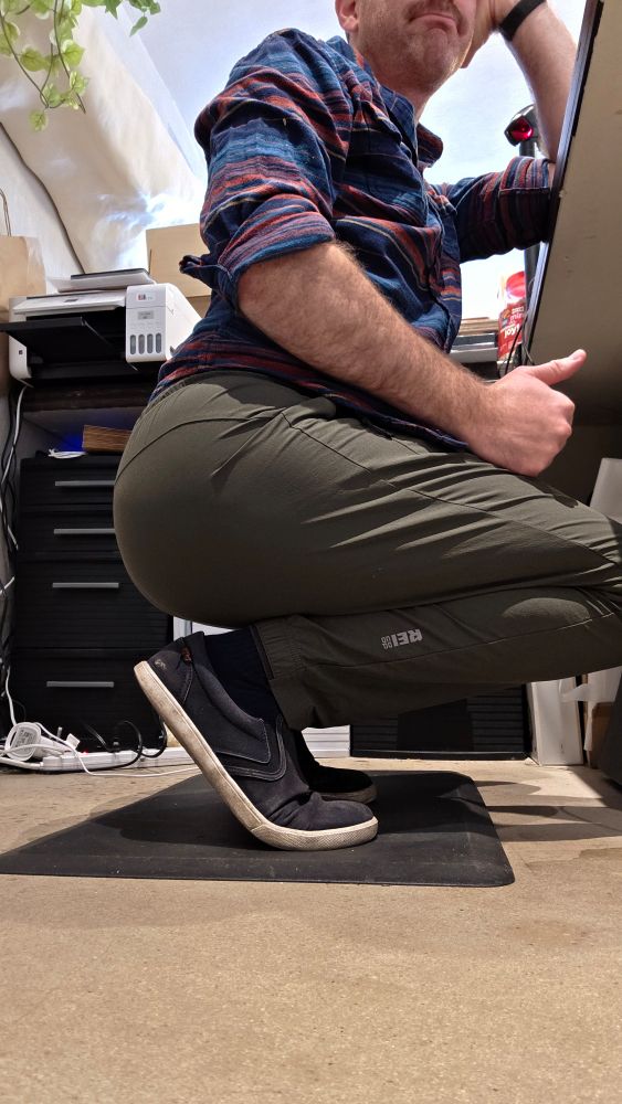 Me squatting at a low counter at work in green pants, plaid shirt, with a little thumbs up, and a big ol fat ass