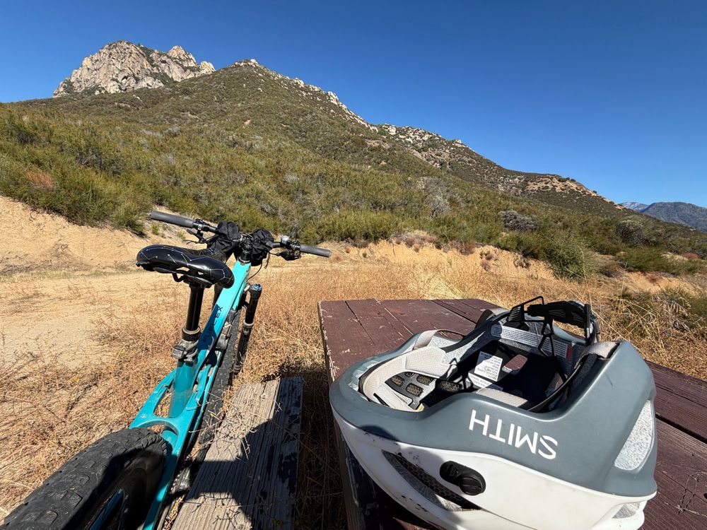 The same blue mountain bike leaning against a brown picnic table with the white helmet on the picnic table in the very near foreground. In the background are brown grass and green hills with some rocks and blue sky