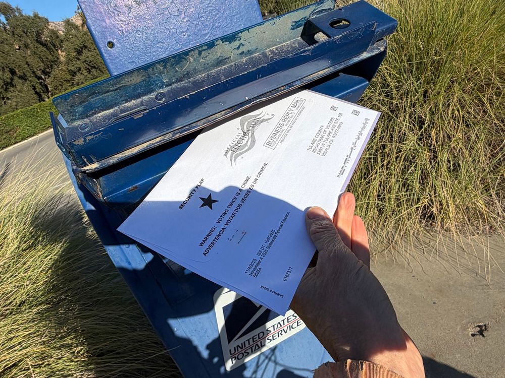 A hand putting a ballot addressed to the Tulare County Registrar of Voters into a mailbox