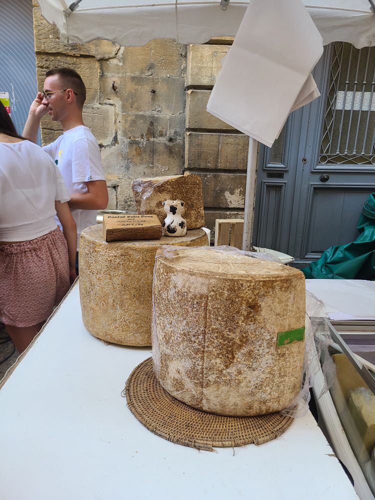 Two huge rounds of cheese sitting on a table in front of a stone building. The cheeses are as large as small stools.