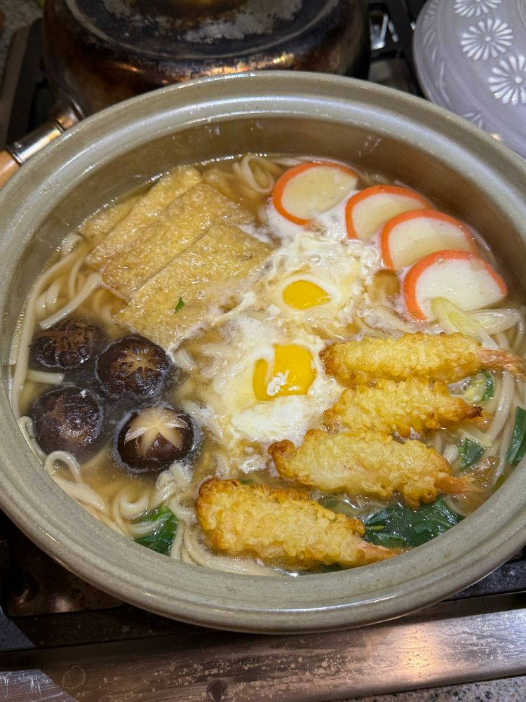Nabeyaki udon: udon noodles cooked in a clay pot. Garnished with slices of fried tofu, kamaboko, shrimp tempura, shiitake, two eggs poached in the broth, and some dandelion greens