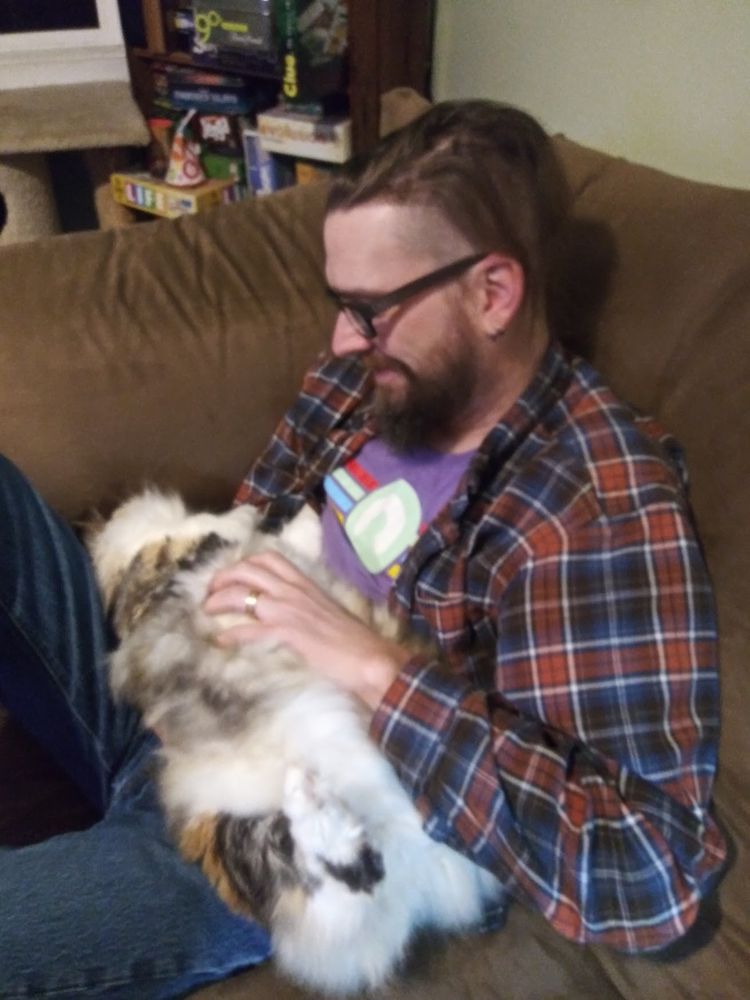 Middle aged guy with glasses in a flannel shirt, sitting on a couch. In his lap is an incredibly fluffy cat, laying on her back while he rubs her tummy.