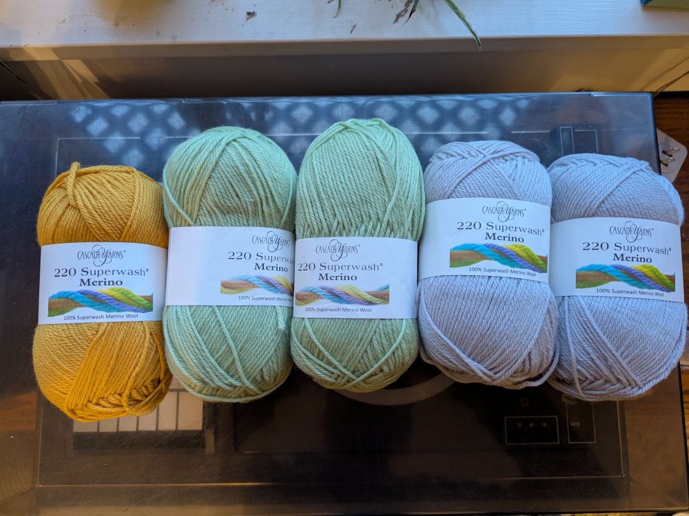 Five skeins of yarn, still in their packaging, labeled "Cascade Yarns Super wash Merino". One is orange, two are soft green, two are soft grey/purple