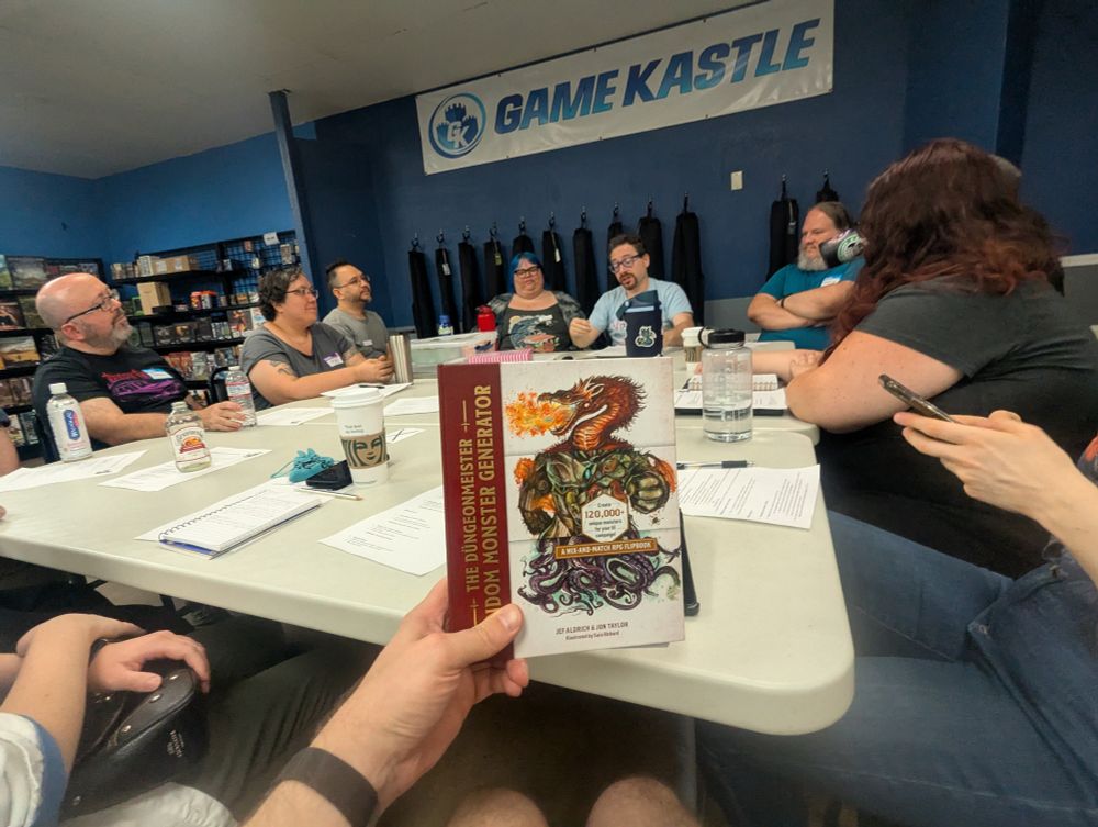A group of people sit around a table in a Game Kastle with papers and drinks in front of them. In the foreground is a copy of the Dungeonmeister Random Monster Generator.